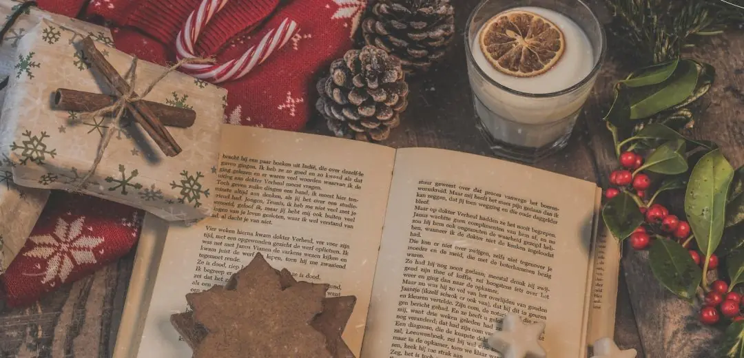 A cozy Christmas scene with an open book on a wooden table, surrounded by wrapped gifts tied with cinnamon sticks, red-and-white candy canes, pinecones, a glass of milk with a dried orange slice on top, holly with red berries, a red winter sweater with snowflake patterns, and festive cookies shaped like stars.