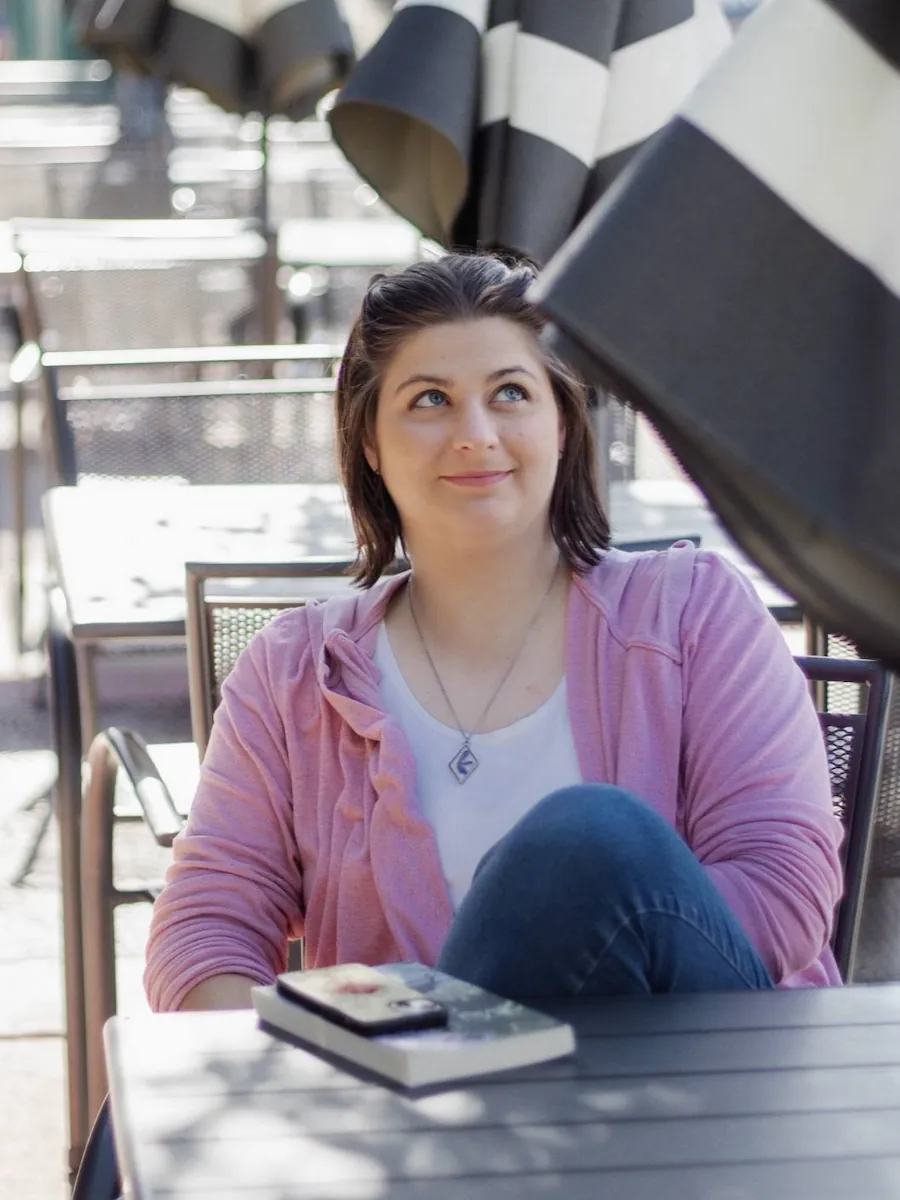 Mandy sitting at a table on the sidewalk outside a cafe. She's wearing a pink cardigan, white shirt, jeans, and a silver necklace. Her phone and 'My World of Glass' lay on the table.